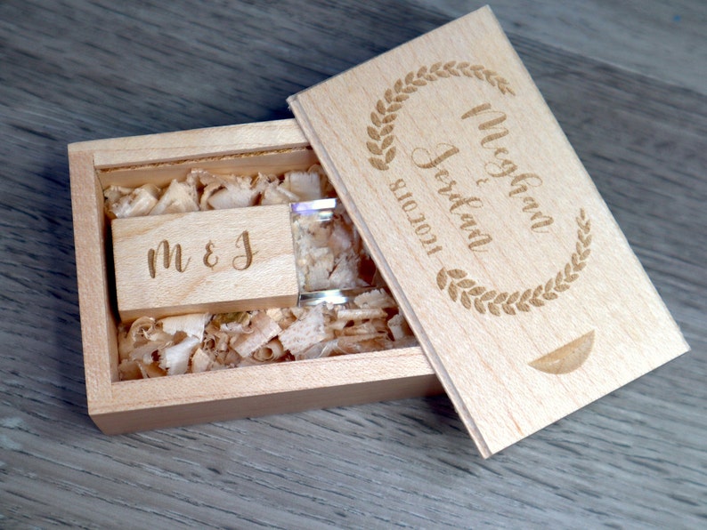 K&ouml;nnte beinhalten: Ein h&ouml;lzernes USB-Flash-Laufwerk und Box-Set. Der Deckel der Box ist mit den Namen "Meghan & Jordan" und dem Datum "8.10.2021" in einem Lorbeerkranz graviert. Das USB-Laufwerk ist mit den Initialen "M & J" graviert. Die Box ist mit Holzsp&auml;nen gef&uuml;llt.