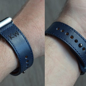 May include: A dark blue leather watch band with white stitching and the initials "KW" engraved on the band. The band is shown on a person's wrist.