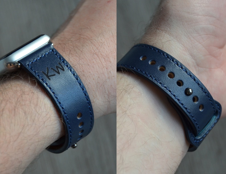 May include: A dark blue leather watch band with white stitching and the initials "KW" engraved on the band. The band is shown on a person's wrist.