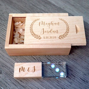 K&ouml;nnte beinhalten: Eine hellfarbene Holzkiste mit Schiebedeckel, graviert mit den Namen "Meghan & Jordan" und dem Datum "8.10.2021" in einem Lorbeerkranz. Ein kleineres Holzst&uuml;ck mit "M & J" liegt neben einem durchsichtigen Kristallblock.