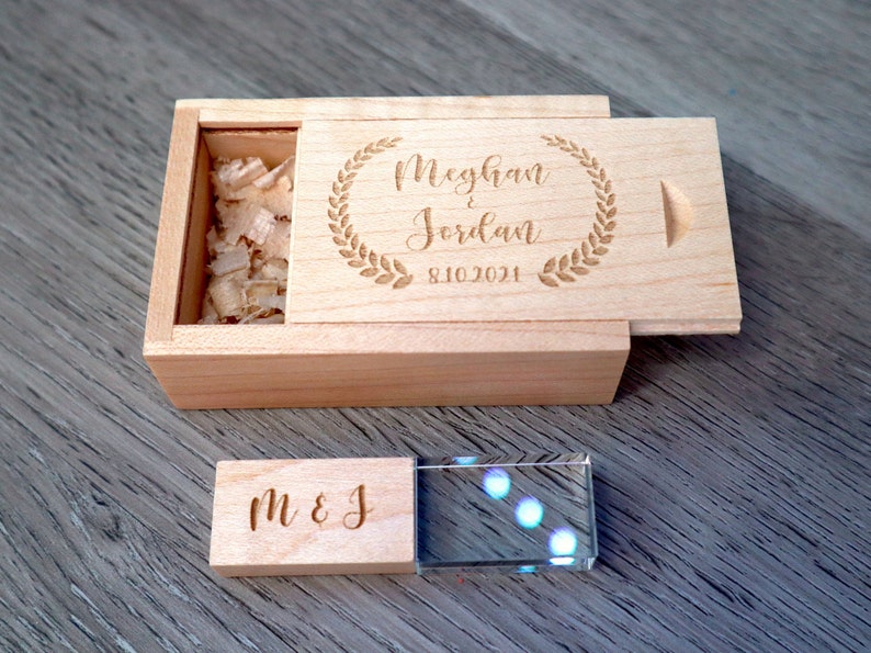 K&ouml;nnte beinhalten: Eine hellfarbene Holzkiste mit Schiebedeckel, graviert mit den Namen "Meghan & Jordan" und dem Datum "8.10.2021" in einem Lorbeerkranz. Ein kleineres Holzst&uuml;ck mit "M & J" liegt neben einem durchsichtigen Kristallblock.