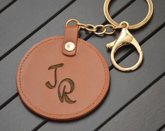 Op de afbeelding: Een bruine leren sleutelhanger met een rond label met de initialen "JR" gegraveerd in een cursief schrift. De sleutelhanger is bevestigd aan een gouden metalen ring en een karabijnhaak.