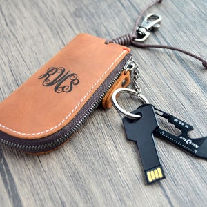 Può includere: Un portachiavi in ​​pelle marrone con una tasca con cerniera. La tasca ha un monogramma "RWS" sul davanti. Il portachiavi ha un anello portachiavi argentato con un'unità USB nera a forma di chiave e un multitool.