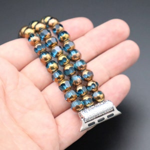 May include: A three-strand beaded Apple Watch band with blue and gold beads. The band has a silver metal clasp with a diamond-like pattern.