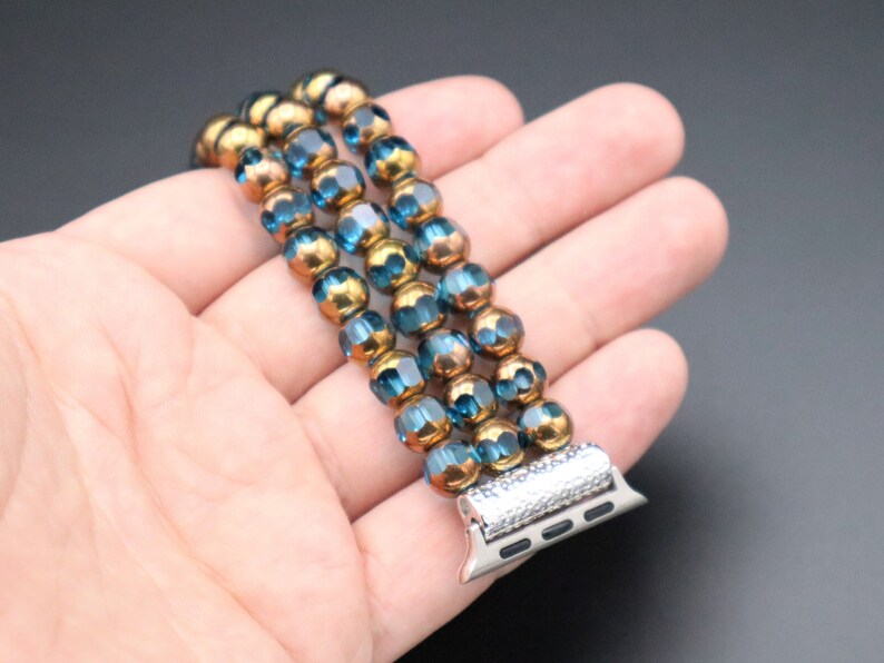 May include: A three-strand beaded Apple Watch band with blue and gold beads. The band has a silver metal clasp with a diamond-like pattern.