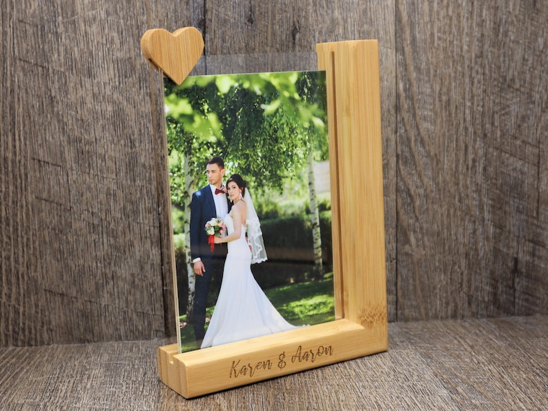 May include: A wooden photo frame with a heart-shaped clip holding a photograph of a couple. The frame is made of light-colored wood and has the names "Karen & Aaron" engraved on the base. The photo shows a bride and groom.
