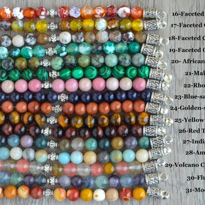 May include: A close-up of 15 rows of different colored gemstone beads with silver clasps. The beads are labeled with their names, including Fire Agate, Olive Agate, Coffee Agate, Green Agate, African Turquoise, Malachite, Rhodonite, Blue-sand Stone, Golden-sand Stone, Yellow Tiger's Eye, Red Tiger's Eye, Indian Agate, Amazonite, Volcano Cherry Quartz, Fluorite, and Mookaite.