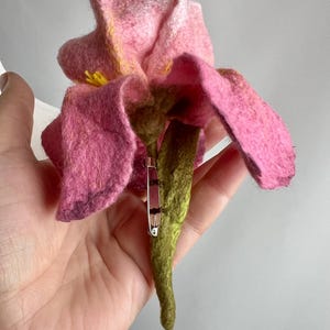 May include: A handmade felt flower brooch in shades of pink, yellow, and green. The brooch features a detailed flower design with layered petals and a green stem. A silver safety pin is attached to the back for securing the brooch.