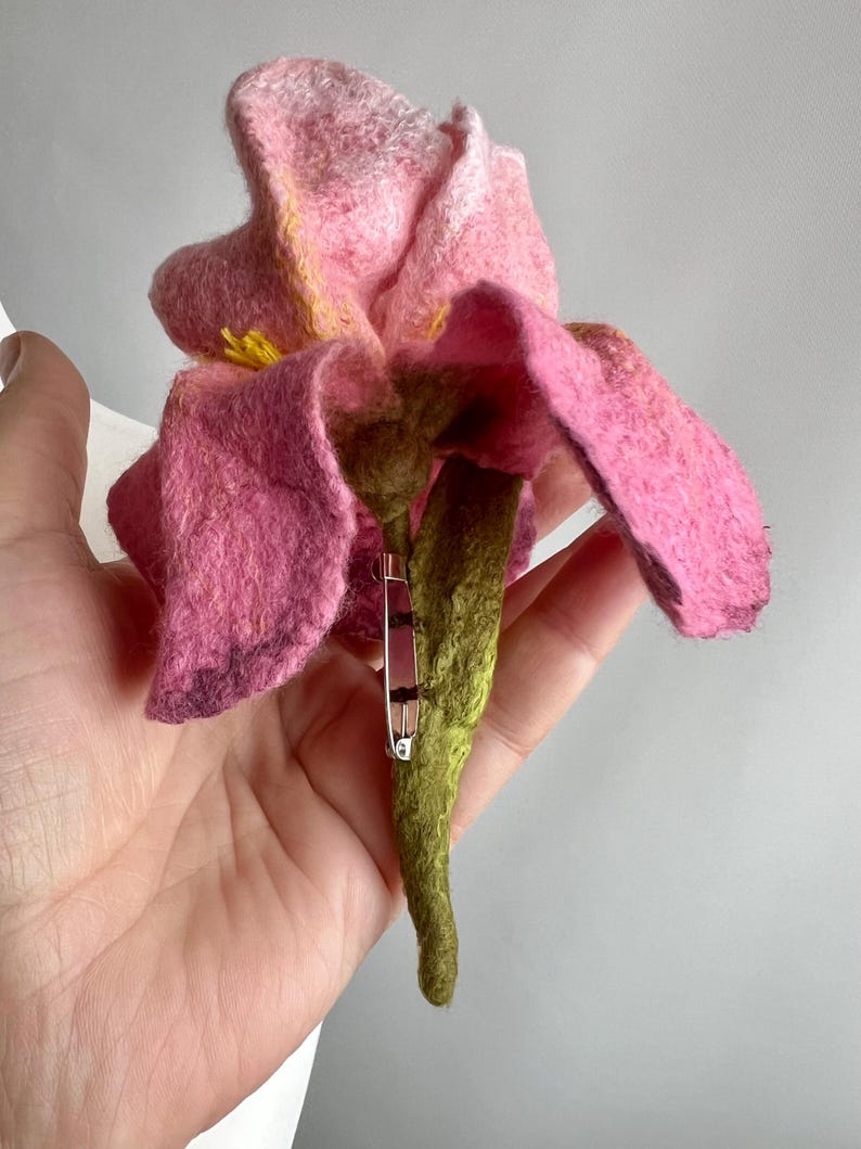May include: A handmade felt flower brooch in shades of pink, yellow, and green. The brooch features a detailed flower design with layered petals and a green stem. A silver safety pin is attached to the back for securing the brooch.
