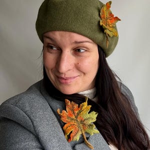 May include: An olive green beret with a felted maple leaf accent. A matching maple leaf brooch is pinned to a gray blazer. The leaves feature shades of orange, yellow, and green.