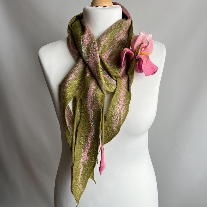 May include: A handmade scarf in olive green and pink, draped on a mannequin. The scarf features vertical stripes of pink and olive green. A pink flower is attached to the scarf, adding a decorative touch. The scarf is made of a soft, textured material.