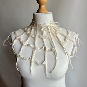 Pode incluir: Um colarinho de crochê na cor creme com um padrão aberto semelhante a uma teia de aranha, com bordas irregulares e pendentes. O colarinho é preso na frente com uma fita de cetim fina na cor creme, amarrada em um laço, sobre um torso de manequim branco.