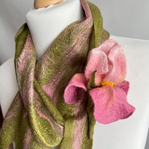 May include: A felt scarf in shades of olive green and pink, draped over a mannequin. A large, pink felt flower with yellow accents is attached to the scarf, adding a floral accent. The scarf's texture is visible.