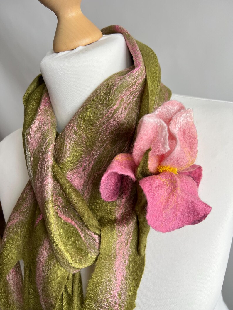 May include: A felt scarf in shades of olive green and pink, draped over a mannequin. A large, pink felt flower with yellow accents is attached to the scarf, adding a floral accent. The scarf's texture is visible.