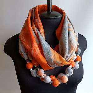 May include: A vibrant orange and grey scarf is displayed on a black mannequin. The scarf features a soft texture and a striking pattern. A necklace with orange and grey felt balls and clear beads is draped around the scarf.
