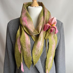 May include: A handmade scarf with a floral design. The scarf is olive green with pink accents and a large pink flower attached. The scarf is draped over a gray blazer on a mannequin. The scarf is made of a soft, textured material.