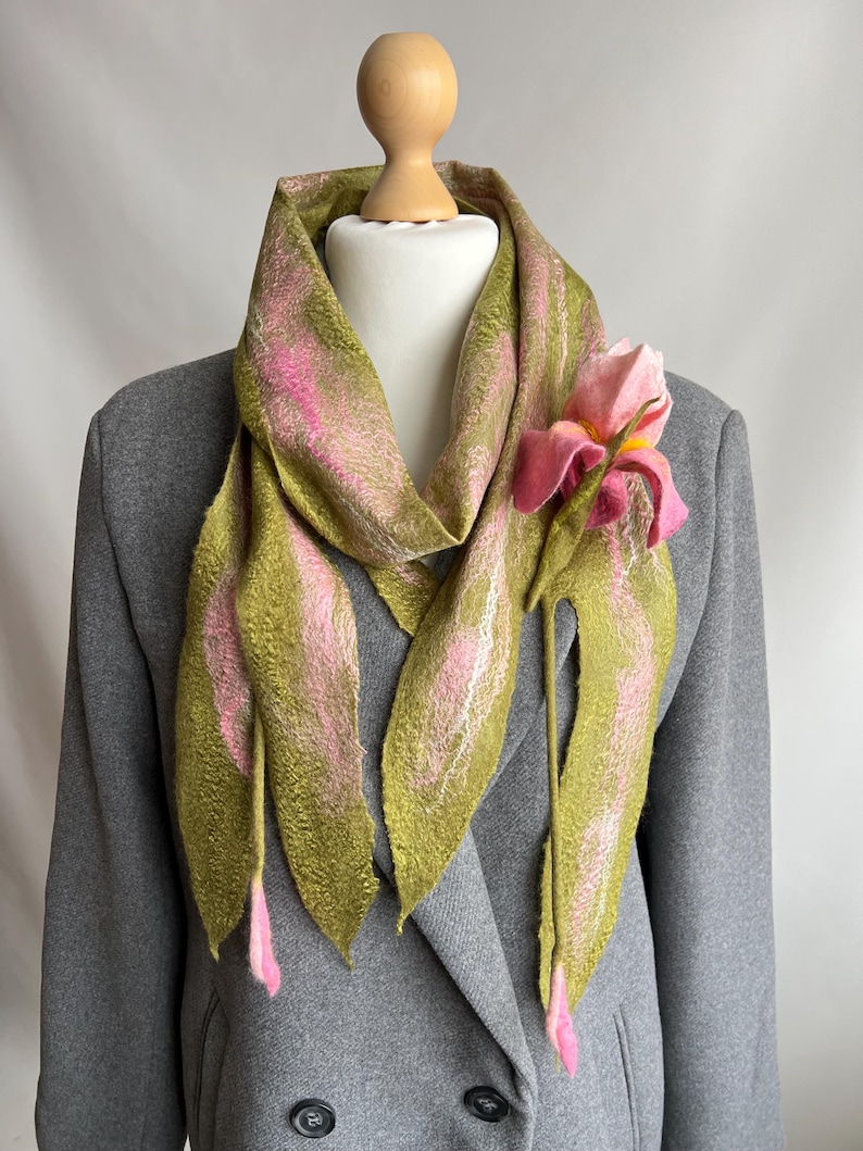 May include: A handmade scarf with a floral design. The scarf is olive green with pink accents and a large pink flower attached. The scarf is draped over a gray blazer on a mannequin. The scarf is made of a soft, textured material.