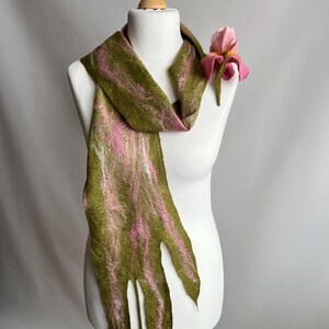 May include: A handmade scarf in olive green with pink and white streaks, displayed on a mannequin. The scarf has a unique, fringed edge and is adorned with a pink felt flower. The scarf is a unique accessory.