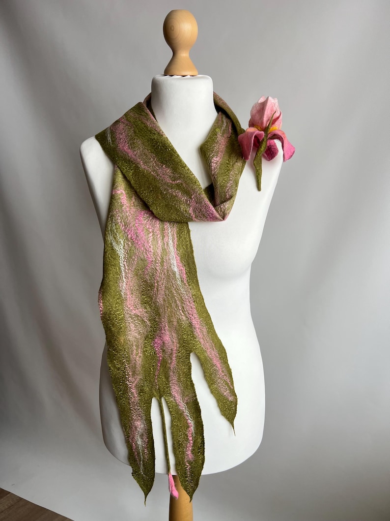 May include: A handmade scarf in olive green with pink and white streaks, displayed on a mannequin. The scarf has a unique, fringed edge and is adorned with a pink felt flower. The scarf is a unique accessory.