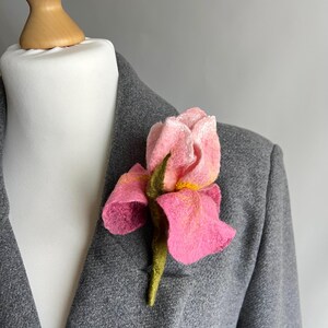 May include: A pink felt flower brooch pinned to a gray blazer. The flower has a detailed design with yellow accents and a green stem. The blazer is displayed on a white mannequin.