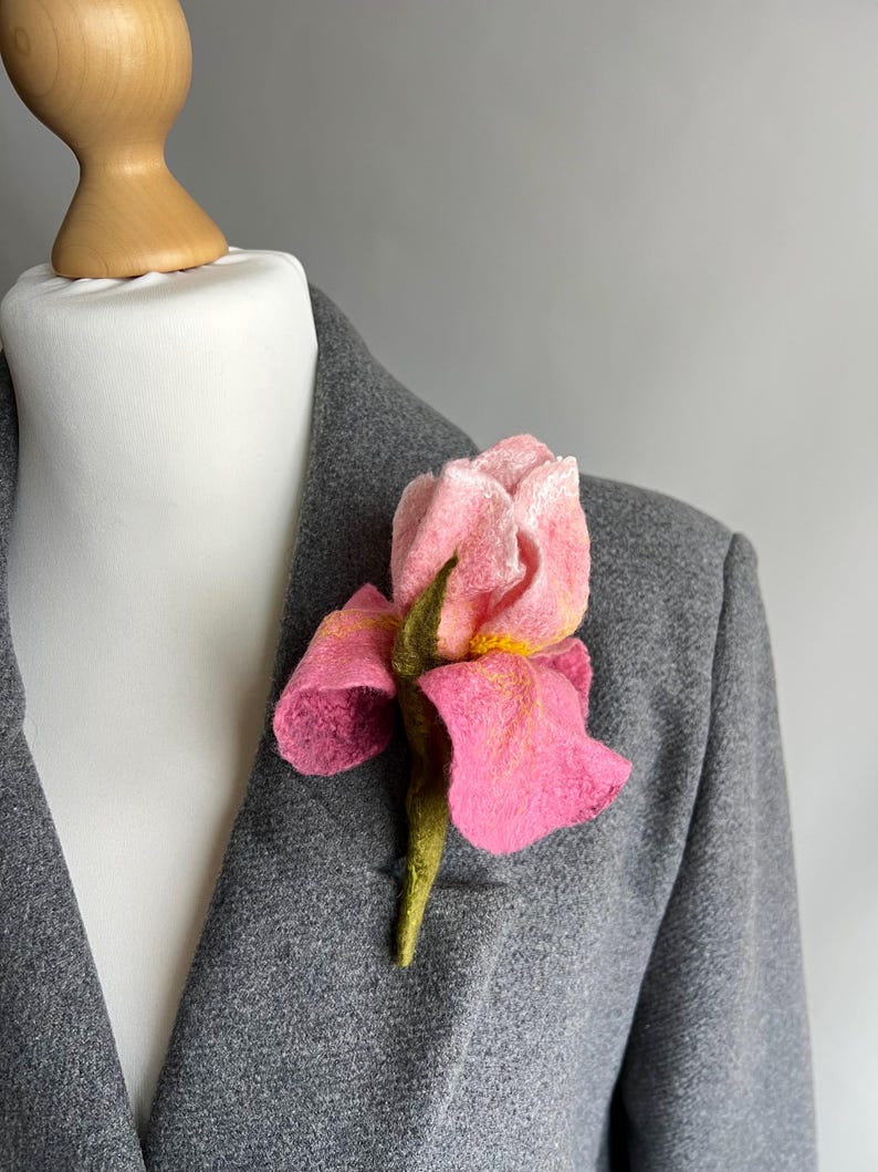 May include: A pink felt flower brooch pinned to a gray blazer. The flower has a detailed design with yellow accents and a green stem. The blazer is displayed on a white mannequin.