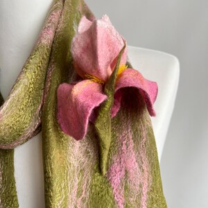 May include: A handmade scarf in shades of green and pink, draped over a mannequin. The scarf features a large, felted pink flower with yellow accents. The scarf's texture is visible, with a blend of colors.