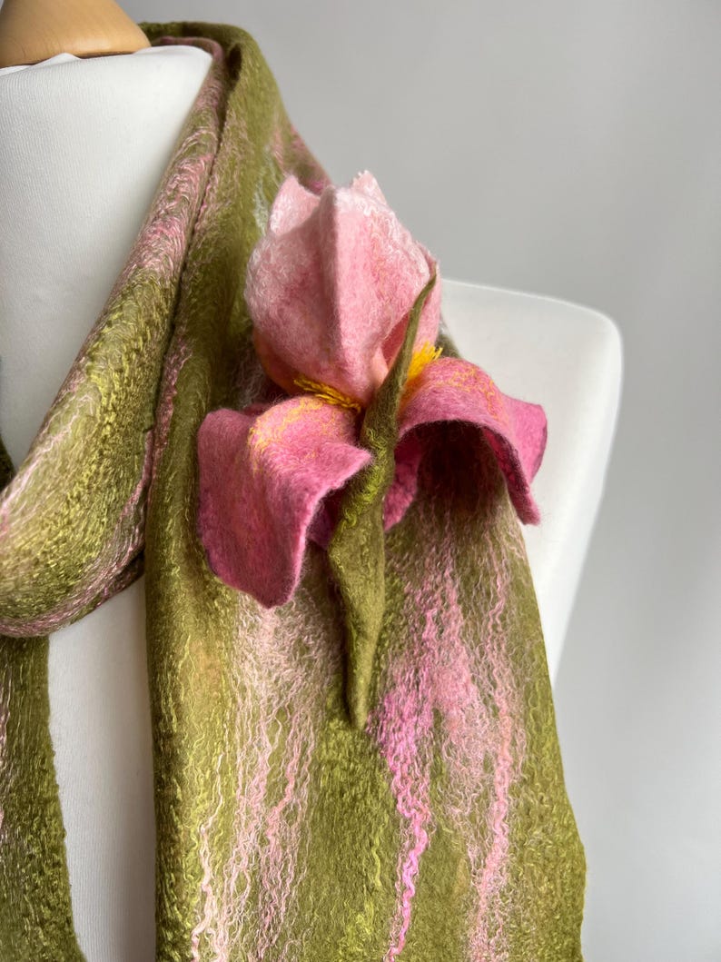 May include: A handmade scarf in shades of green and pink, draped over a mannequin. The scarf features a large, felted pink flower with yellow accents. The scarf's texture is visible, with a blend of colors.