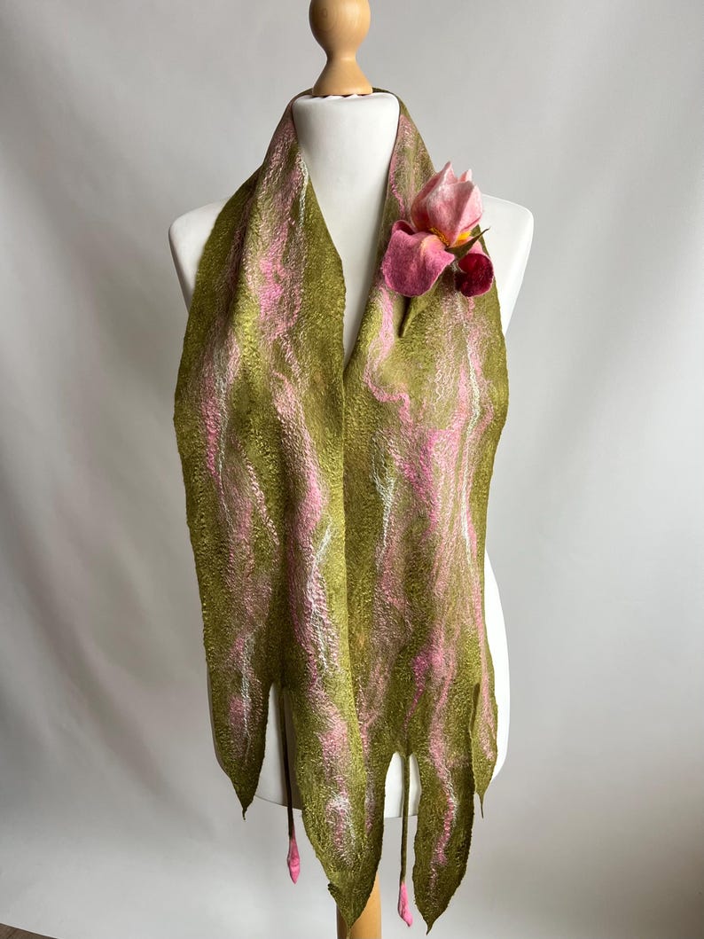 May include: A handmade felt scarf in olive green with pink and white swirls, adorned with a pink felt flower. The scarf has a unique, fringed bottom edge with pink accents, displayed on a mannequin.