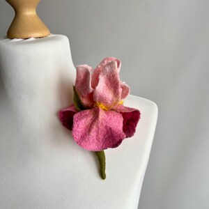 May include: A pink felt flower brooch with a yellow center and green stem. The brooch is displayed on a white mannequin. The flower has multiple layers of pink petals and a textured appearance.