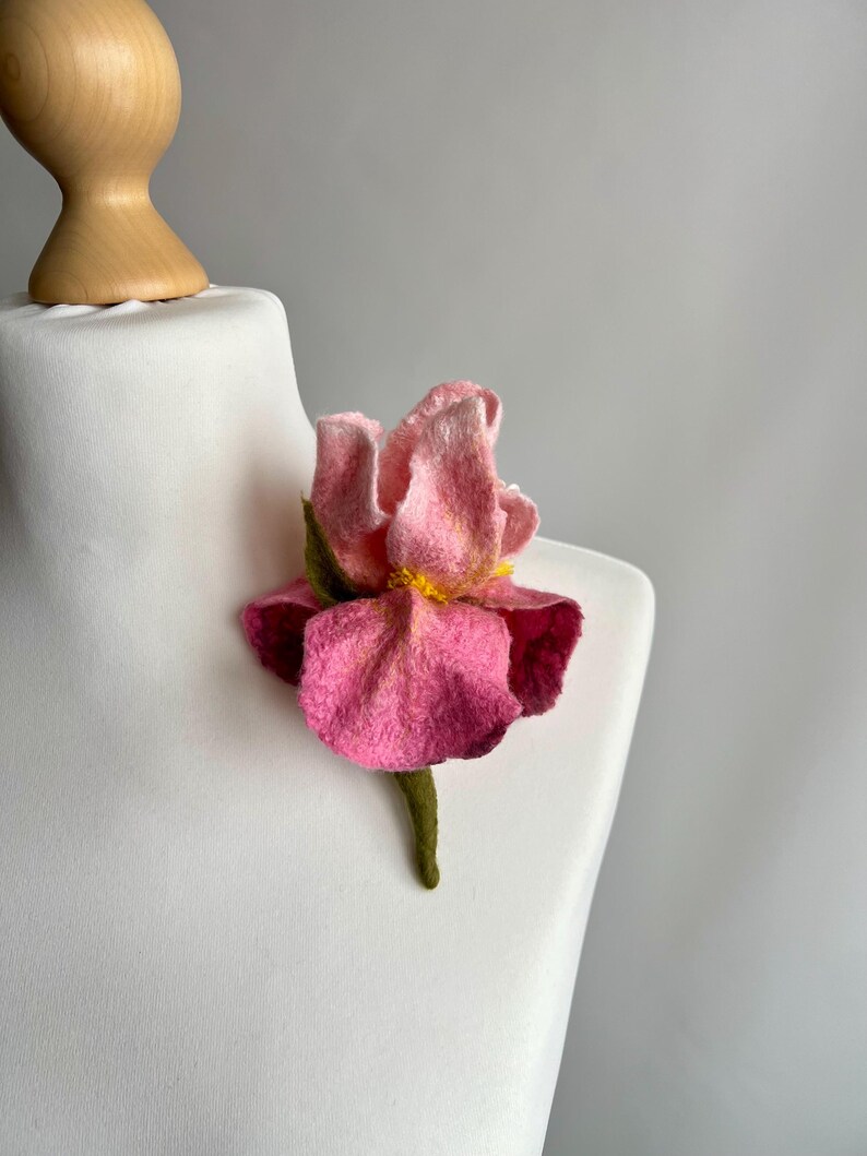 May include: A pink felt flower brooch with a yellow center and green stem. The brooch is displayed on a white mannequin. The flower has multiple layers of pink petals and a textured appearance.