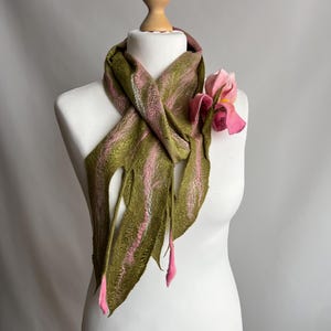 May include: A handmade scarf displayed on a mannequin. The scarf is olive green with pink stripes and fringed edges. A pink flower is attached to the scarf. The scarf is made of felted wool.