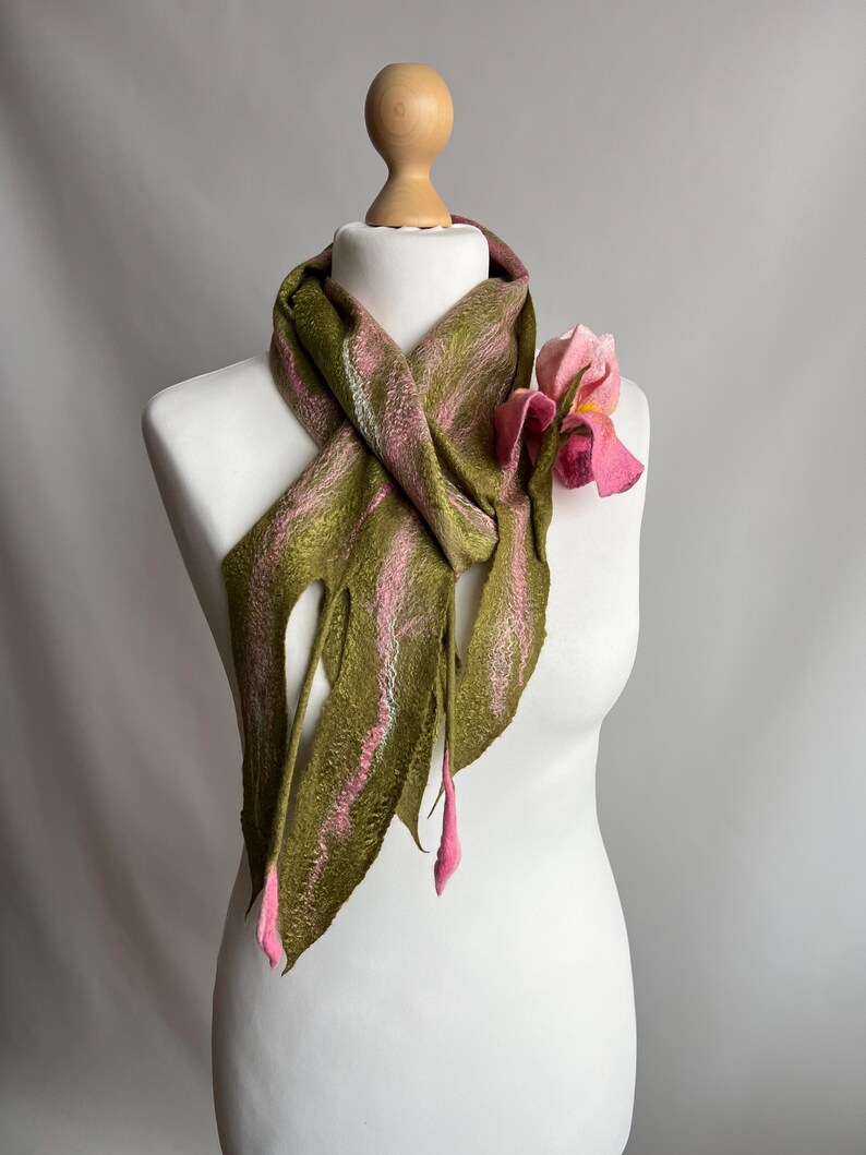 May include: A handmade scarf displayed on a mannequin. The scarf is olive green with pink stripes and fringed edges. A pink flower is attached to the scarf. The scarf is made of felted wool.