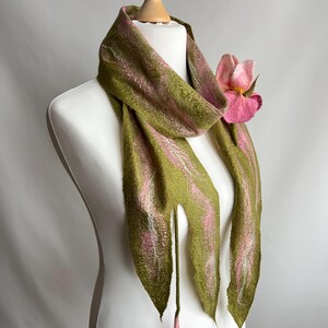 May include: A handmade scarf displayed on a mannequin. The scarf is olive green with pink accents and a large pink flower. The scarf has a unique, textured appearance and is draped to showcase its design.