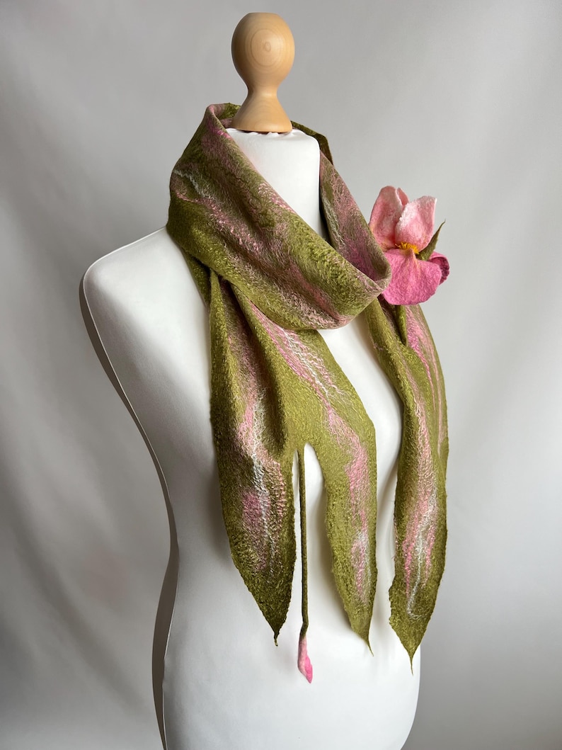 May include: A handmade scarf displayed on a mannequin. The scarf is olive green with pink accents and a large pink flower. The scarf has a unique, textured appearance and is draped to showcase its design.