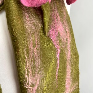 May include: A handmade felt scarf with a floral design. The scarf features a base of olive green with pink accents and a large, three-dimensional pink flower. The texture of the felt is visible, adding to the handcrafted aesthetic.