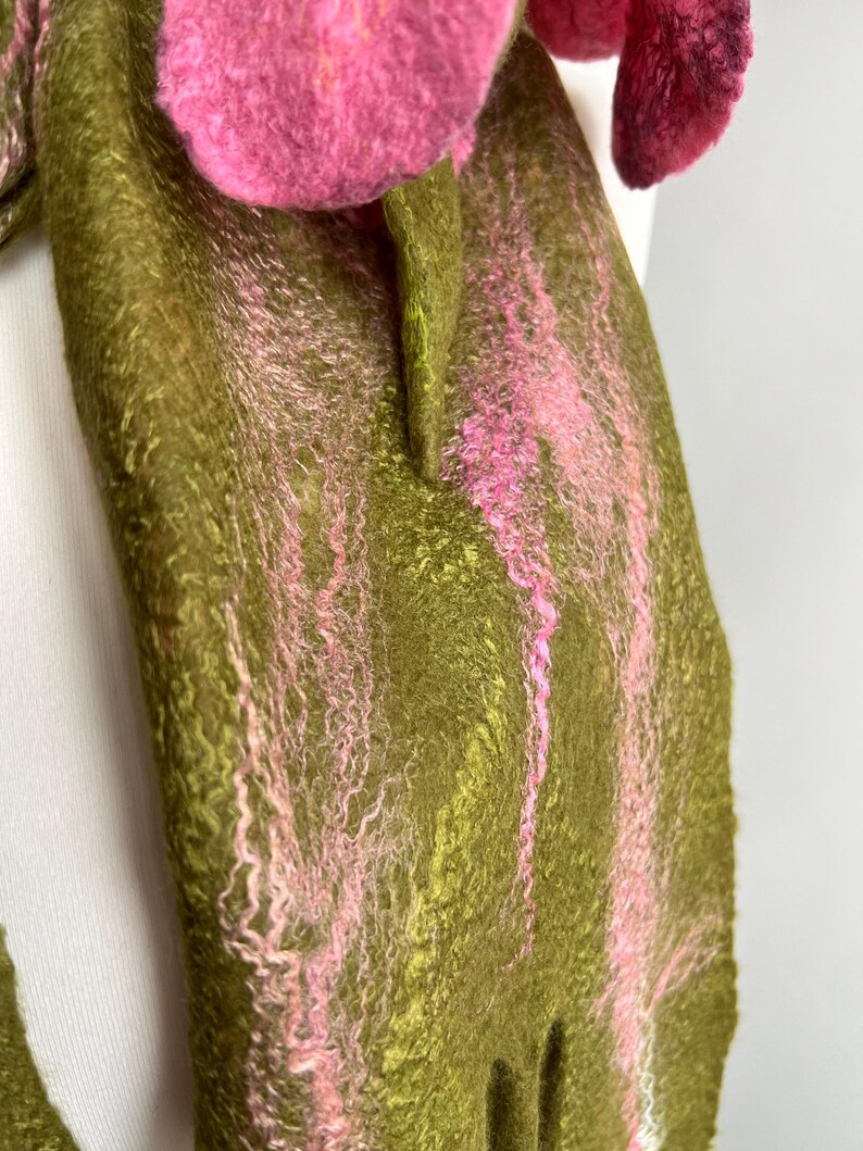 May include: A handmade felt scarf with a floral design. The scarf features a base of olive green with pink accents and a large, three-dimensional pink flower. The texture of the felt is visible, adding to the handcrafted aesthetic.