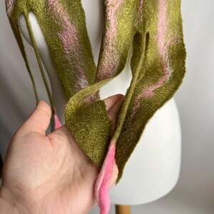May include: Olive green and pink felt scarf. The scarf has a textured appearance with pink stripes running along its length. The ends of the scarf are pointed and finished with pink accents. The scarf is displayed on a white mannequin head.