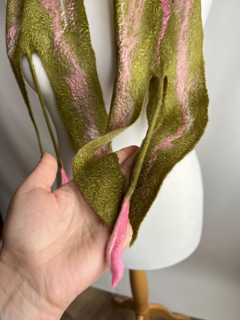 May include: Olive green and pink felt scarf. The scarf has a textured appearance with pink stripes running along its length. The ends of the scarf are pointed and finished with pink accents. The scarf is displayed on a white mannequin head.