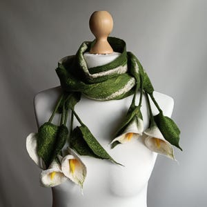 Può includere: Una sciarpa di feltro verde e bianca con fiori di calla bianchi attaccati. La sciarpa è esposta su un manichino bianco.