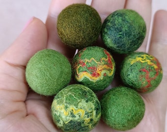 May include: A collection of seven green felt balls, some with yellow and red accents. The balls are in a hand, ready to be used for crafting or decoration.