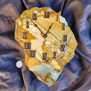 May include: A wall clock crafted from a slab of yellow, tan, and cream-colored jasper stone with a natural, irregular edge. The clock face features dark brown Roman numerals and matching clock hands. A small silver coin is placed for scale.