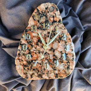 May include: A unique, freeform clock made from a speckled granite stone. The clock face features silver-colored numerals and hands, with a gold-colored center. The stone has a mix of red, black, and white colors.