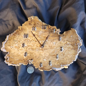 May include: A wall clock crafted from a natural, irregular slice of light brown and cream-colored stone with a swirling pattern. The clock features dark metal hands and raised metallic numerals. A silver coin is placed below for scale.