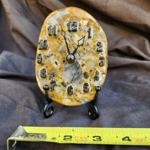 May include: An oval-shaped clock made from a polished, speckled yellow and gray stone. It features raised black numerals from 1 to 12 and black metal hands. The clock rests on a black metal stand, measuring approximately 4 inches in width.
