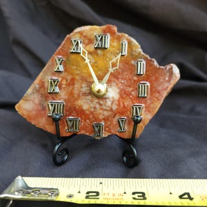 May include: A clock made from a slice of orange and white agate stone, featuring silver Roman numerals and gold-colored hands. The clock rests on a black metal stand, and a yellow measuring tape shows it is approximately 4.5 inches wide.