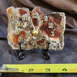 May include: A rectangular clock made from a slice of raw, multi-colored agate stone with deep red, brown, and grey tones. It features raised gold-colored numerals and ornate gold hands. The clock rests on a black metal stand, with a yellow measuring tape showing a width of approximately 4.5 inches.