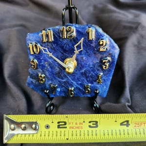 May include: A blue stone clock with gold-colored numbers and hands, mounted on a black metal stand. A yellow measuring tape is positioned in front, showing the clock face is approximately 4 inches wide at its base.