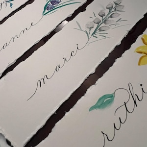 Op de afbeelding: Waterverf naamkaartjes met bloemmotieven en handgeschreven namen. Elke kaart toont een andere bloem, waaronder roze, paarse en gele bloesems, met namen als "Anne" en "Marci" in sierlijke letters.