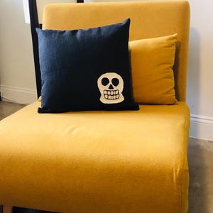 May include: A dark navy blue square pillow with a white skull design, resting on a yellow chair. A smaller yellow pillow is behind it. The chair has wooden legs and is on a woven rug.
