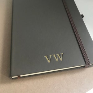 May include: A dark grey leather-bound journal with a gold elastic closure and the initials "VV" embossed in gold on the front cover.